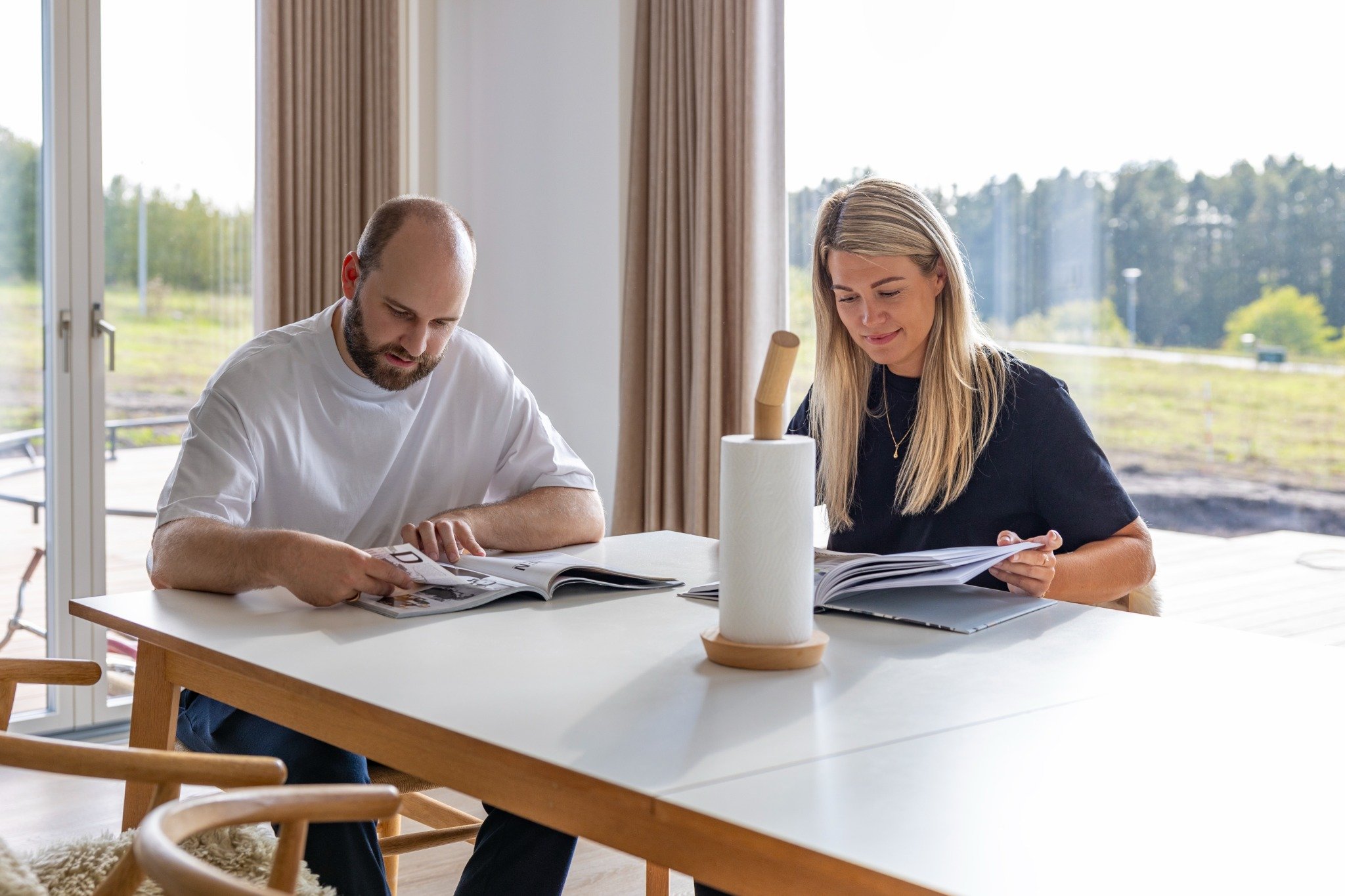 En mand og en kvinde sidder ved et spisebord og bladrer i magasiner i et lyst rum med store vinduer. Udsigten udenfor viser træer og åbent landskab.