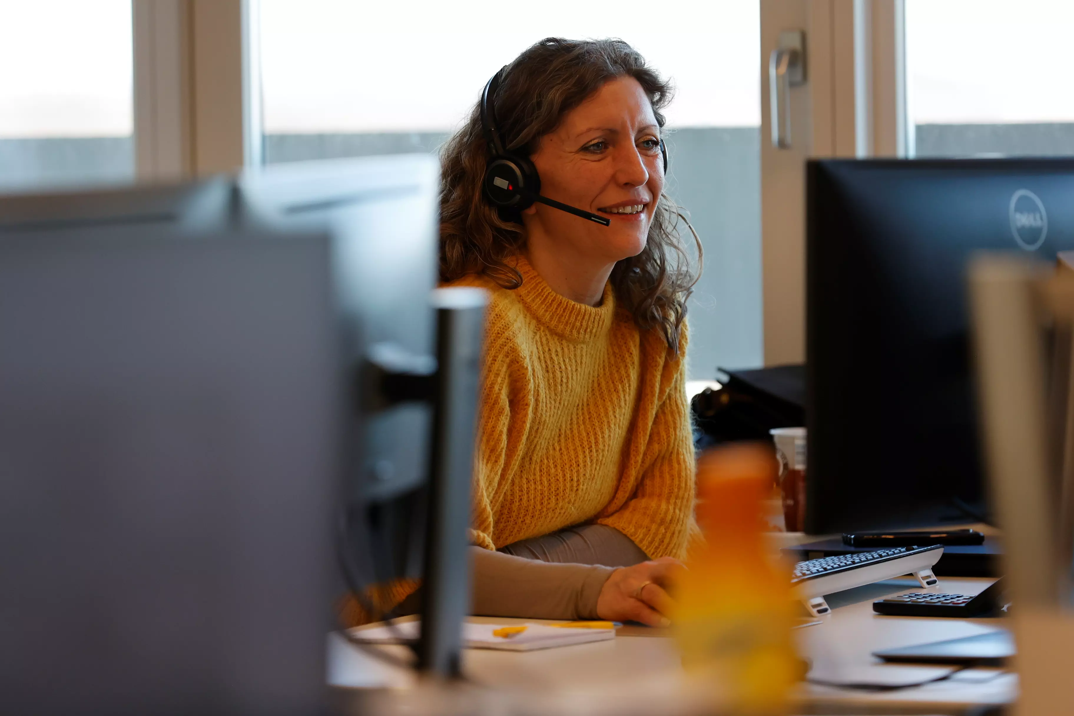 En kvinde sidder og arbejder ved sin computer. Hun kigger ind i skærmen med headset på og smiler.