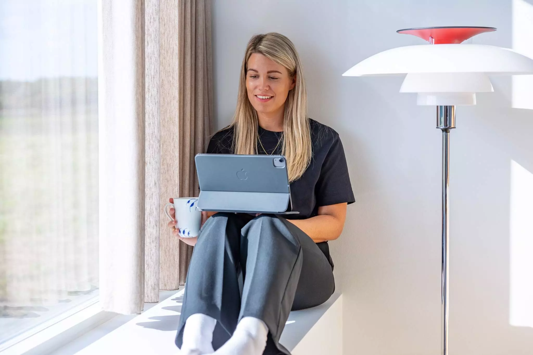 En kvinde sidder afslappet på en vindueskarm med benene trukket op, mens hun holder en tablet i den ene hånd og en kaffekop i den anden. Hun smiler og kigger på tabletten. Sollys strømmer ind gennem vinduet ved siden af hende.