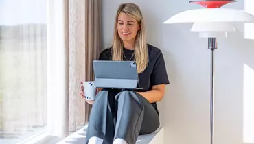 En kvinde sidder afslappet på en vindueskarm med benene trukket op, mens hun holder en tablet i den ene hånd og en kaffekop i den anden. Hun smiler og kigger på tabletten. Sollys strømmer ind gennem vinduet ved siden af hende.