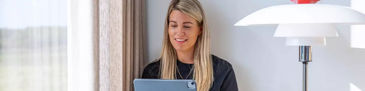 En kvinde sidder afslappet på en vindueskarm med benene trukket op, mens hun holder en tablet i den ene hånd og en kaffekop i den anden. Hun smiler og kigger på tabletten. Sollys strømmer ind gennem vinduet ved siden af hende.