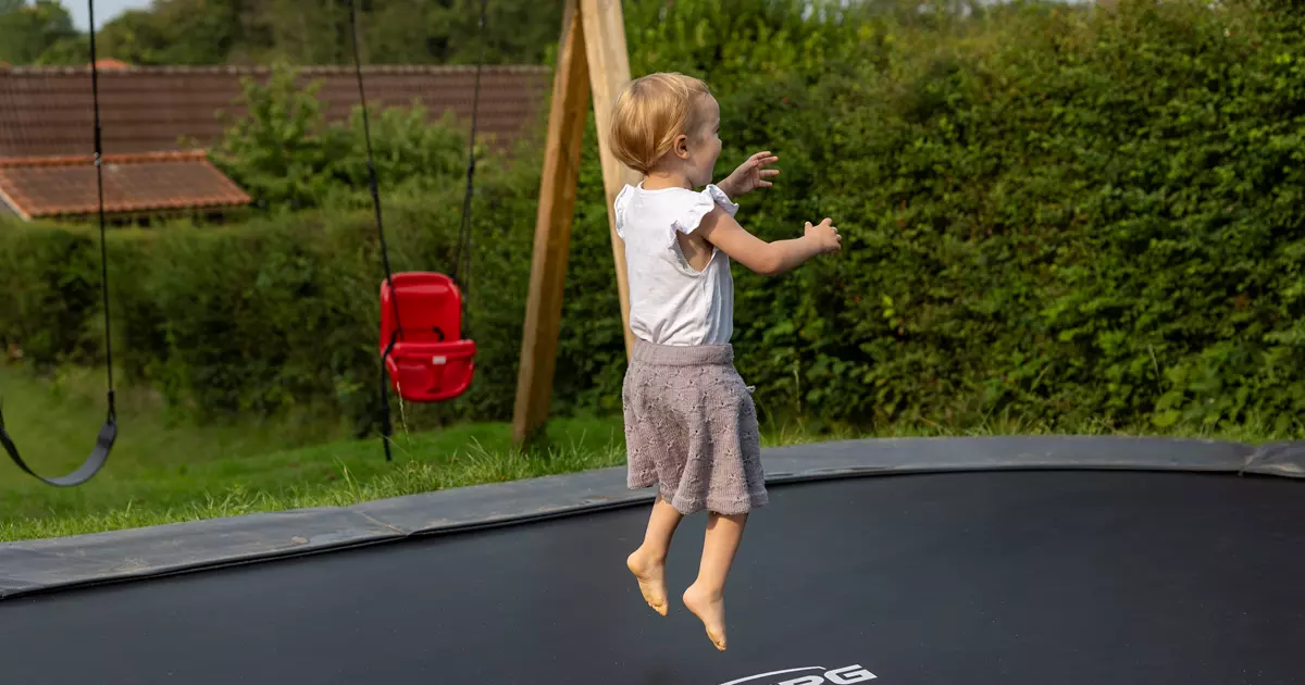Et lille barn hopper på en sort trampolin i en have. I baggrunden ses en rød gynge og grønne buske.