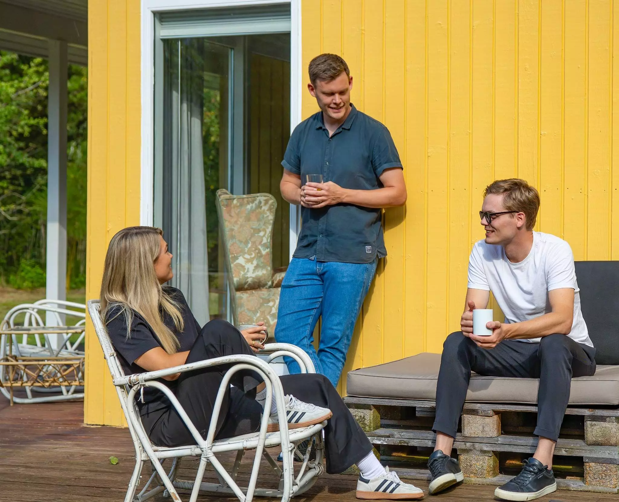 Tre personer sidder og står ved en gul træbygning. En kvinde sidder i en hvid fletstol og taler med to mænd, hvoraf den ene står og holder et glas vand, og den anden sidder på en sofa og holder en kaffekop. De ser ud til at nyde en afslappet samtale udendørs.