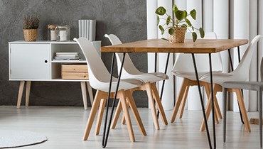 Et moderne spisestueområde med et træbord og hvide stole med træben. På bordet står en potteplante, og i baggrunden ses et hvidt skab med pyntegenstande og en grå væg.