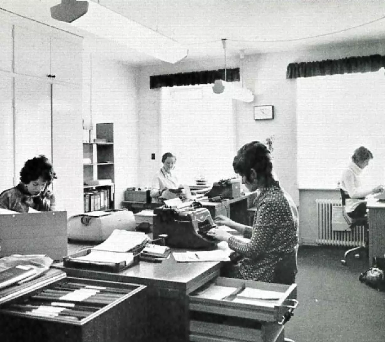 Billedet viser et sort-hvidt kontormiljø fra en tidligere tid, sandsynligvis fra midten af det 20. århundrede. Flere kvinder arbejder ved skriveborde med skrivemaskiner og papirstakke omkring sig. Rummet er lyst op af naturligt lys fra vinduer med gardiner, og der er reoler og mapper i baggrunden, som antyder en administrativ eller sekretærfunktion. Stemningen virker koncentreret og præget af datidens arbejdsrutiner.