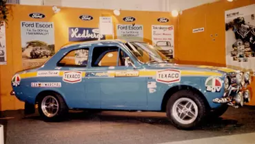 En klassisk blå Ford Escort udstillet på en messe med orange baggrund og Ford-logoer. Bilen er dekoreret med mærkater fra sponsorer som Texaco, AGV, Goodyear og Marchal, samt rally-striber i gult og hvidt. Forlygterne er forstærket til rallybrug, og der er informationsplakater og reklamer om Ford Escort på væggen bag bilen.