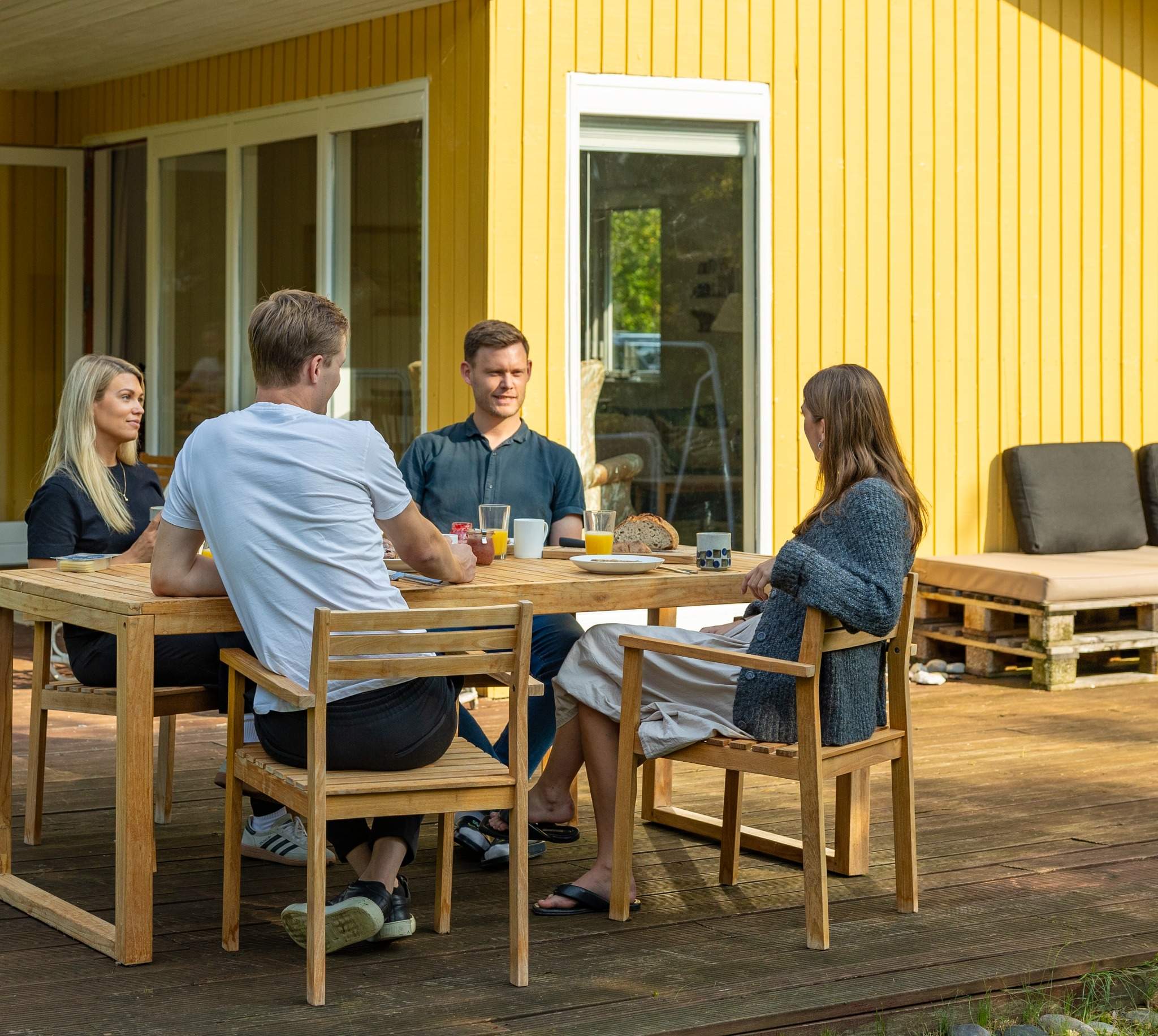 Fire personer sidder udenfor et sommerhus, i et havemøbelsæt. De sidder og slapper af og hygger sig sammen. 