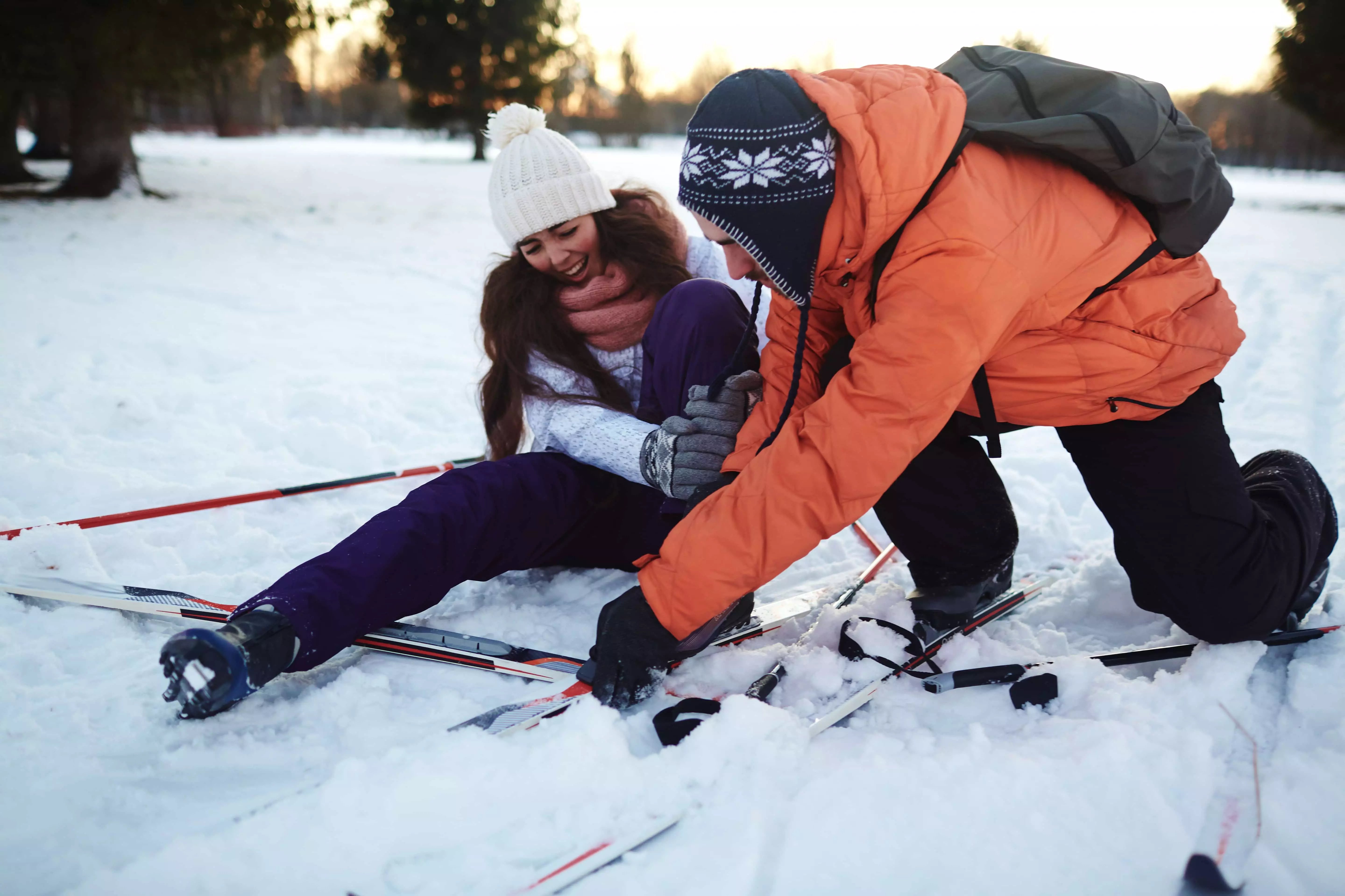 To personer på ski sidder i sneen, hvor en hjælper den anden op. Den ene person bærer en orange jakke og hue med mønster, mens den anden har en hvid hue og lilla skibukser. Baggrunden viser et sneklædt landskab med træer i skumringen.