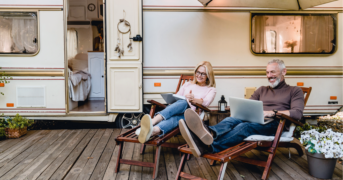 To ældre mennesker, der sidder i udendørs lænestile foran deres campingvogn. Der er god stemning og de hygger sig.