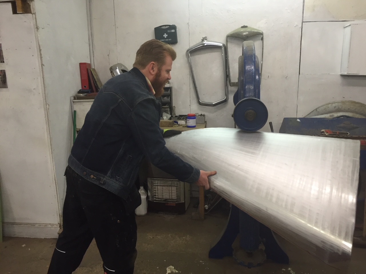  En mand med skæg og en denimjakke arbejder i et værksted, hvor han holder og former et stort stykke metal. I baggrunden ses forskellige værktøjer og udstyr, herunder en blå engelsk hjulmaskine, en spejlet bilgrill og en arbejdsbænk. Manden er fokuseret og ser ud til at bearbejde metallet med præcision.