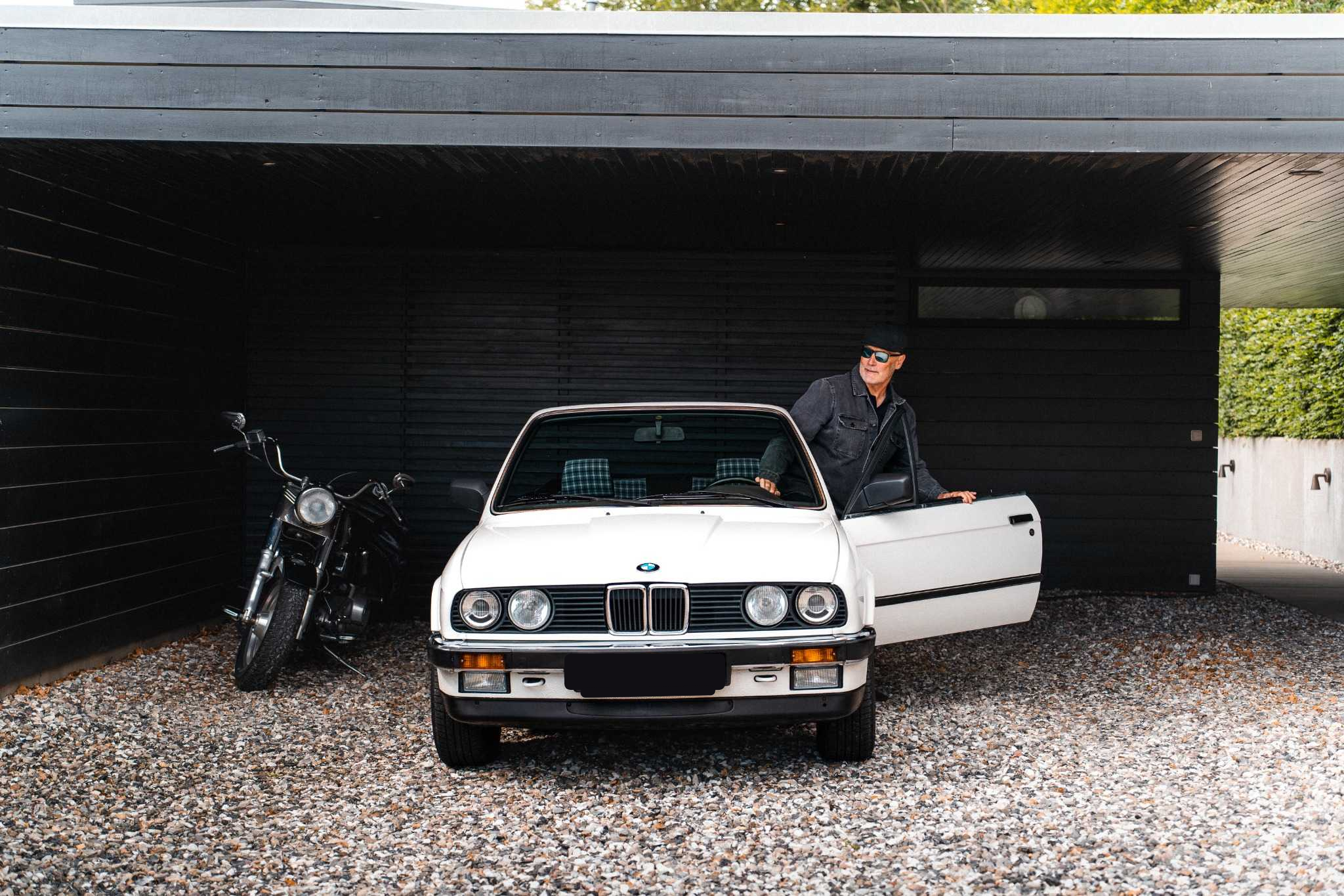 Grøn klassisk Alfa Romeo Spider cabriolet kører på en landevej, med føreren synlig bag rattet. Bilen har åbent tag, og omgivelserne viser en sløret baggrund med marker og træer, hvilket skaber en dynamisk følelse af bevægelse.