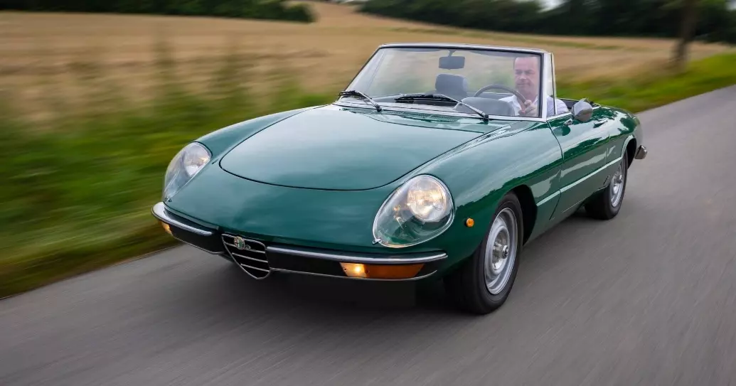 Grøn klassisk Alfa Romeo Spider cabriolet kører på en landevej, med føreren synlig bag rattet. Bilen har åbent tag, og omgivelserne viser en sløret baggrund med marker og træer, hvilket skaber en dynamisk følelse af bevægelse.