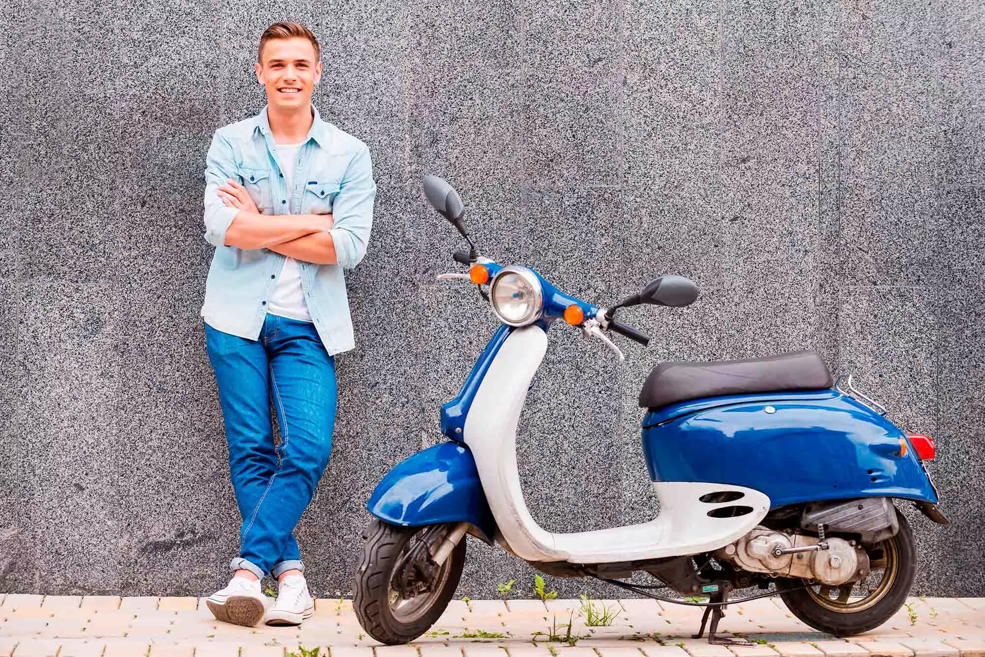 En ung mand i denimjakke og jeans står afslappet med armene over kors ved siden af en blå scooter. Baggrunden er en grå væg, og han smiler til kameraet.