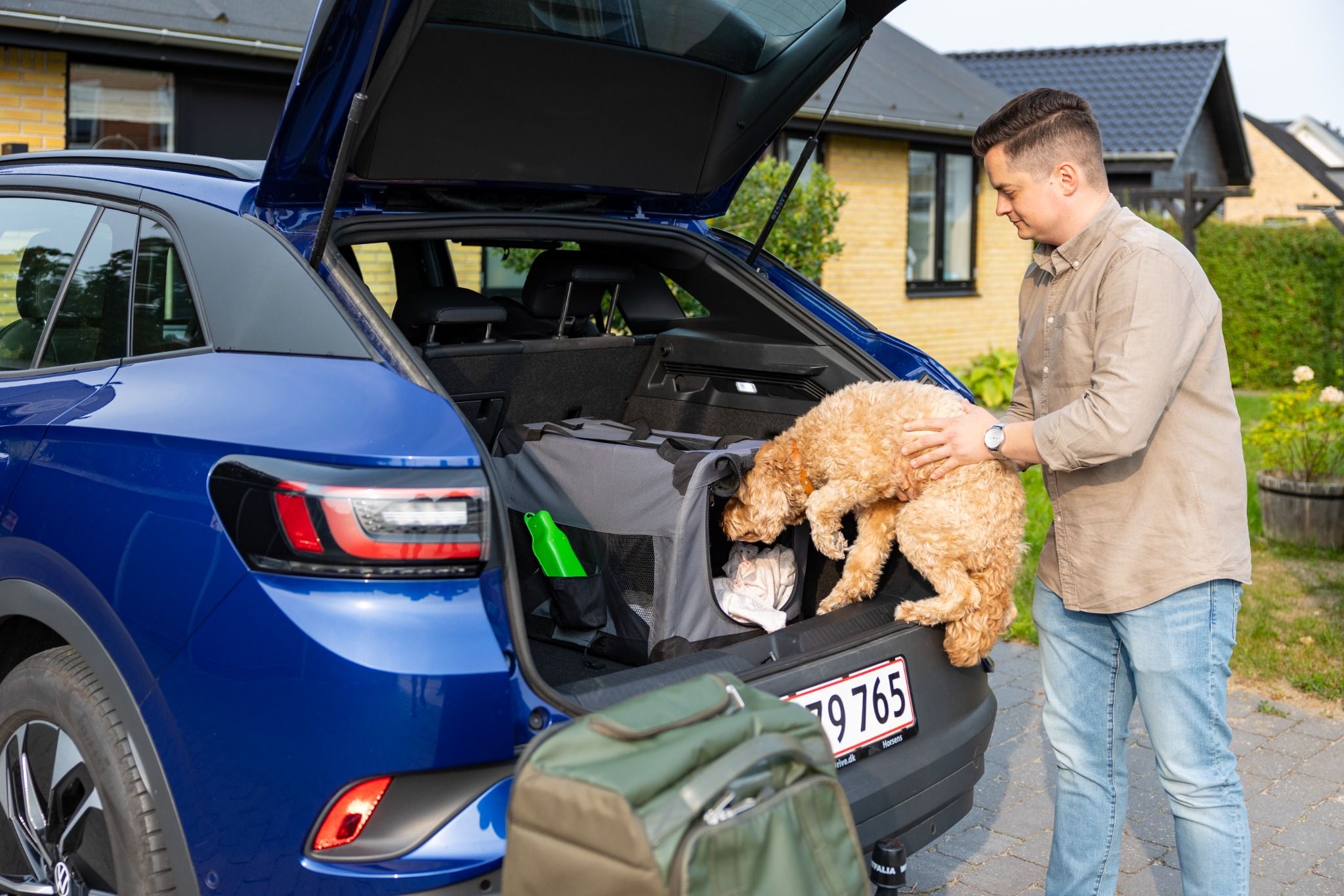 En mand placerer sin hund i en hundetransportkasse i bagagerummet på en blå bil, mens en grøn taske står ved siden af. Baggrunden viser en moderne villa med gul murstensfacade.
