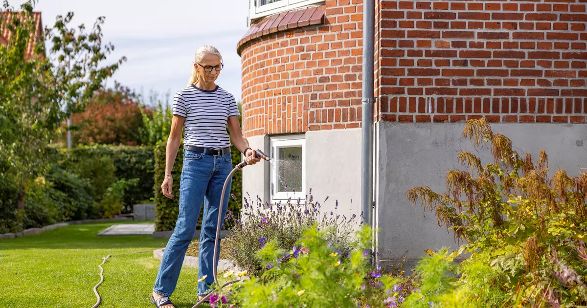 En kvinde i stribet t-shirt og jeans vander et blomsterbed med en haveslange foran en rund murstensbygning. Blomsterbedet er fyldt med farverige planter, og baggrunden viser en velplejet græsplæne og buske.