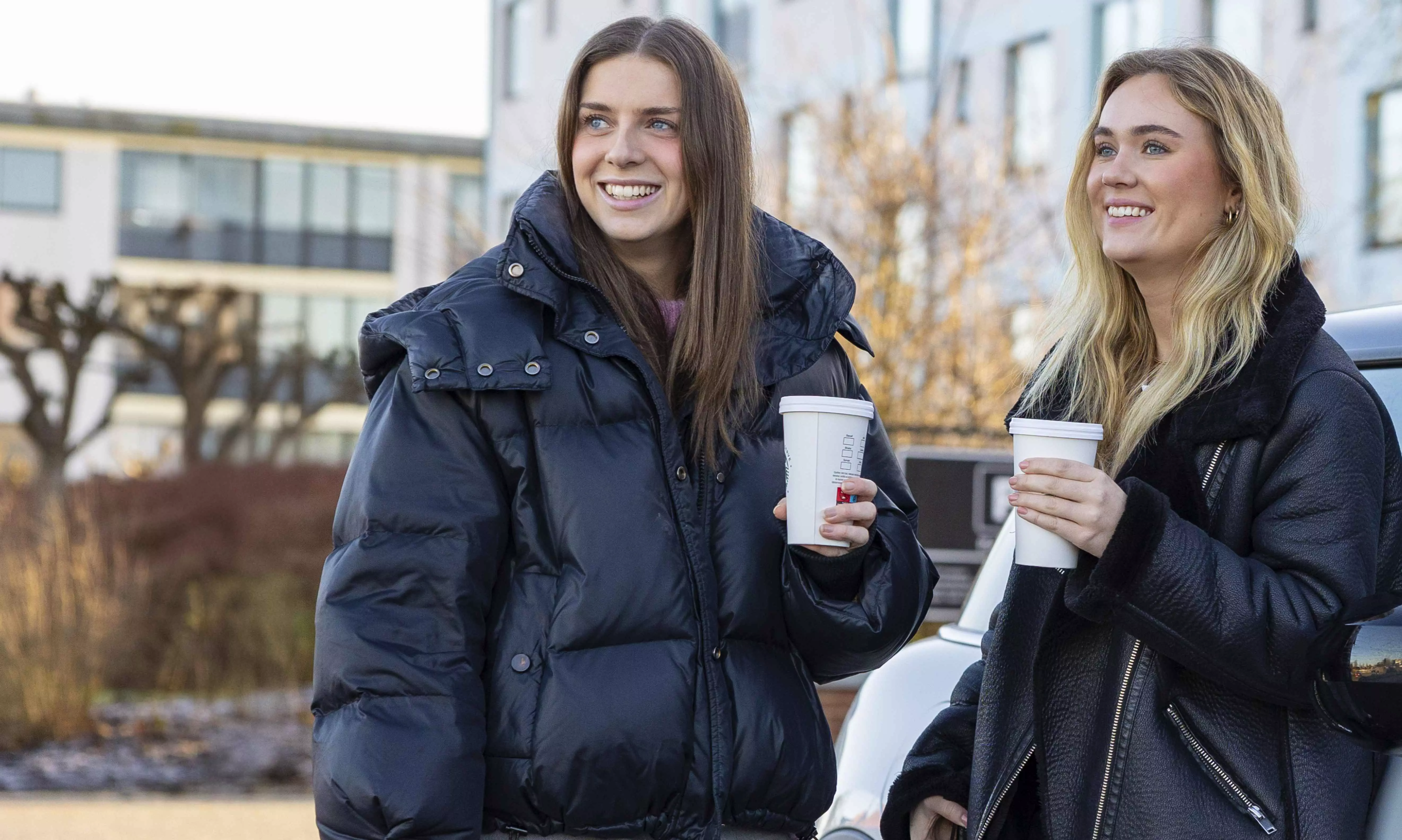 To unge piger står med en kop kaffe i hånden udenfor.