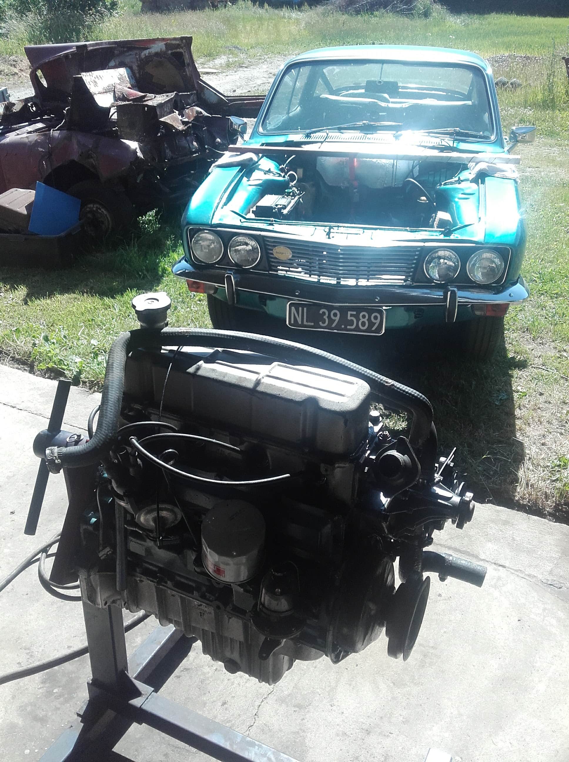 En lyseblå klassisk bil uden motor og med hævet motorhjelm holder udendørs på en græsplæne. Foran bilen ligger en adskilt motor og tilhørende dele på jorden. I baggrunden ses en skadet, rusten bil med knust karosseri og dele, der er skubbet sammen. Græsset er grønt, og solen skinner tydeligt over scenen.