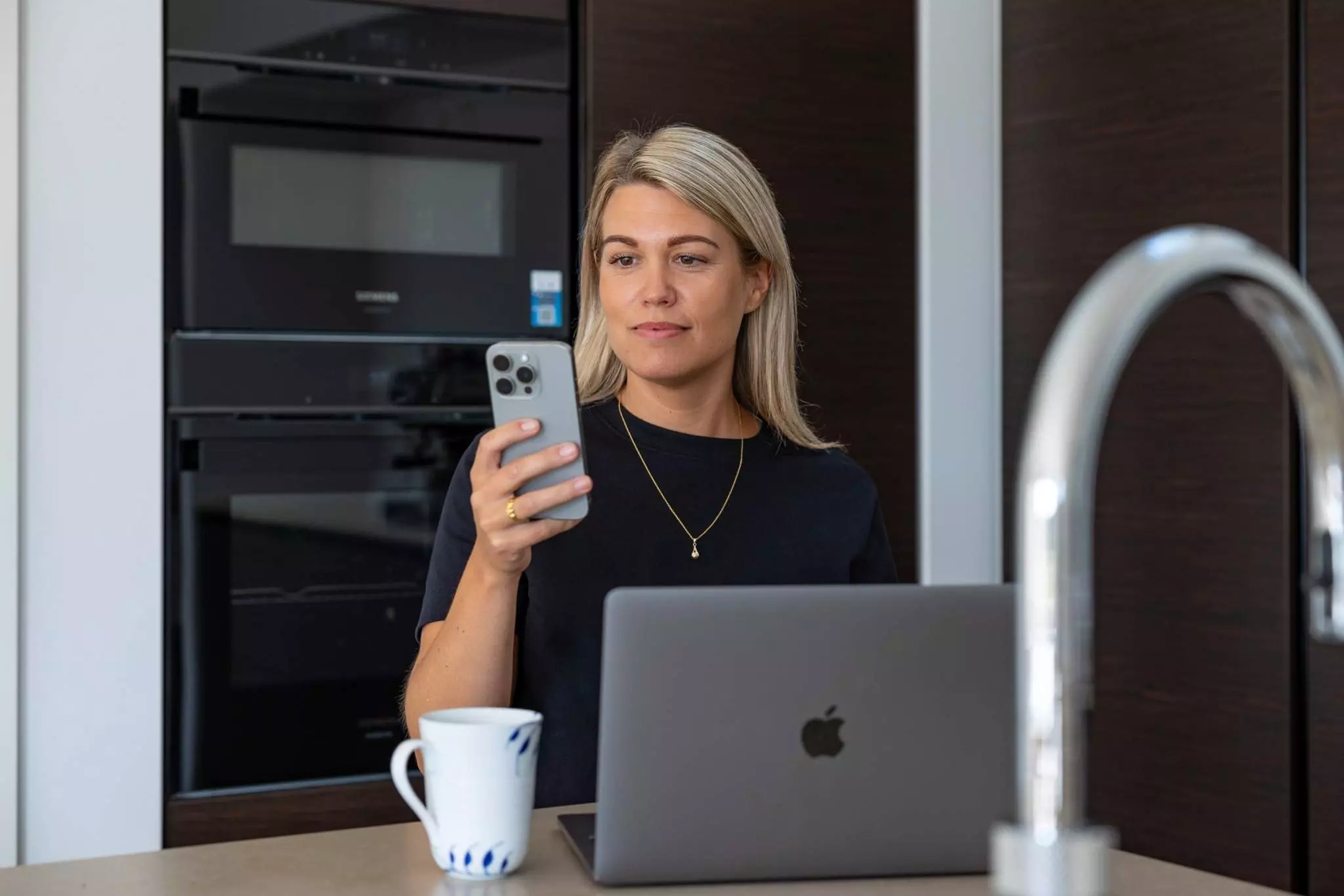 En kvinde sidder ved et køkkenbord med en åben bærbar computer og en kaffekop foran sig. Hun holder en smartphone og kigger koncentreret på skærmen. I baggrunden ses et moderne køkken med en indbygget ovn.