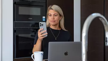 En kvinde sidder ved et køkkenbord med en åben bærbar computer og en kaffekop foran sig. Hun holder en smartphone og kigger koncentreret på skærmen. I baggrunden ses et moderne køkken med en indbygget ovn.