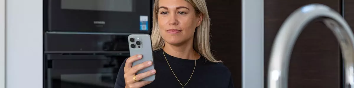 En kvinde sidder ved et køkkenbord med en åben bærbar computer og en kaffekop foran sig. Hun holder en smartphone og kigger koncentreret på skærmen. I baggrunden ses et moderne køkken med en indbygget ovn.