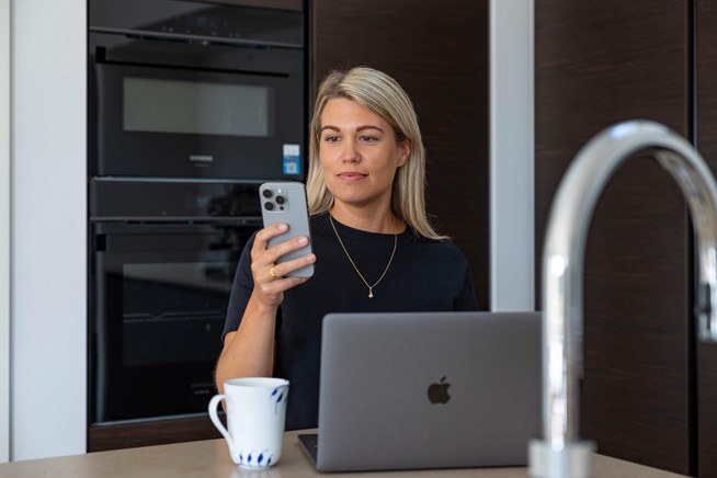 Kvinde bruger smartphone og laptop i køkken. Beskyt din elektronik med en forsikring.