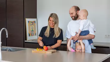 En kvinde skærer frugt på et skærebræt i et moderne køkken, mens hun smiler. Ved siden af står en mand med et spædbarn på armen og et lille barn, der kigger frem bag hans ben. Baggrunden viser mørke køkkenskabe og en farverig illustration.