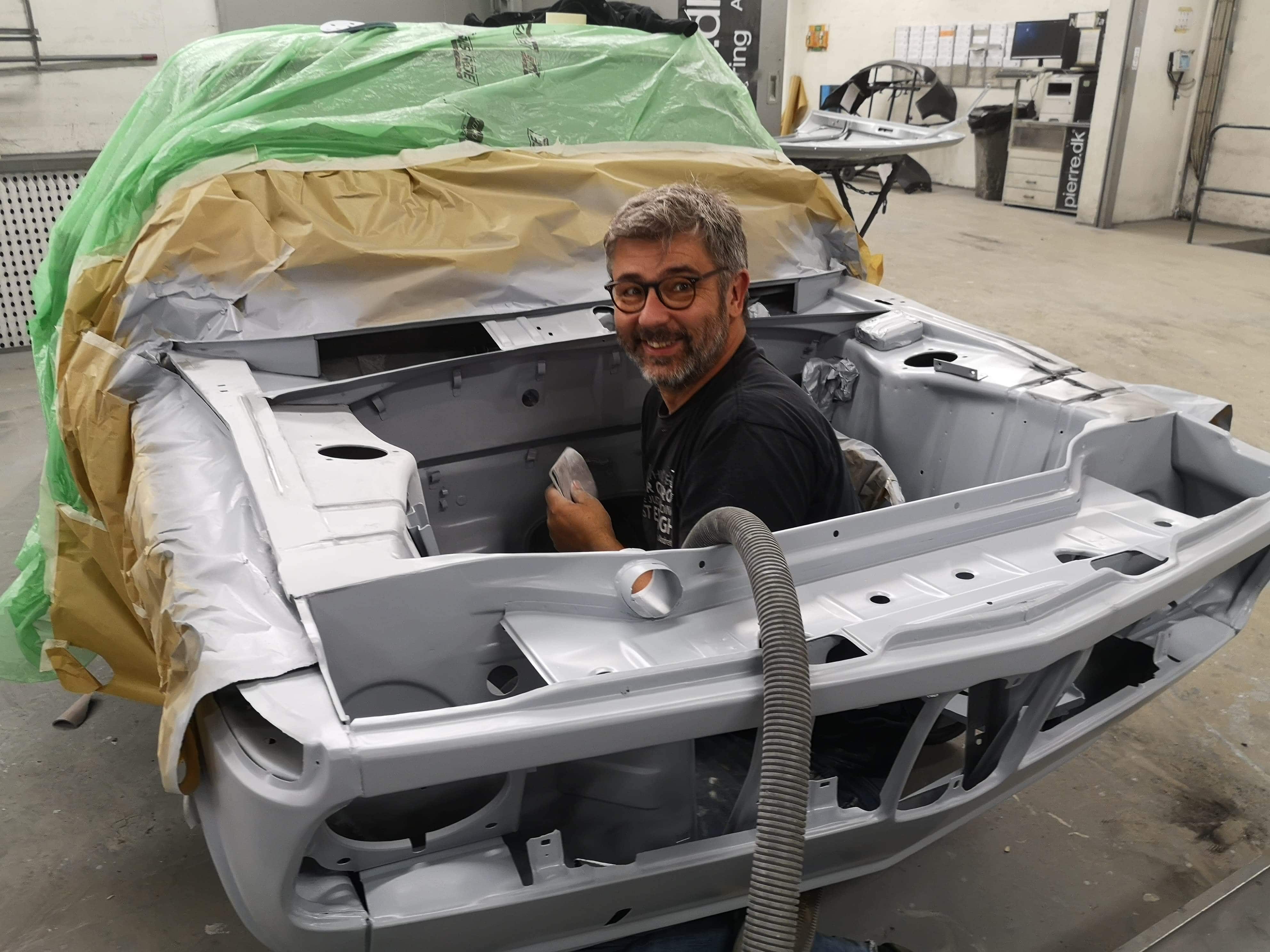 En smilende mand med briller og gråt hår sidder inde i karrosseriet af en bil, der er i gang med at blive restaureret. Bilen er delvist dækket med papir og plastik, og karrosseriet er nylakeret i en sølvgrå farve. Manden arbejder i et professionelt værksted med værktøj og bildele i baggrunden, mens en slange til støvsugning eller ventilation er synlig i forgrunden.