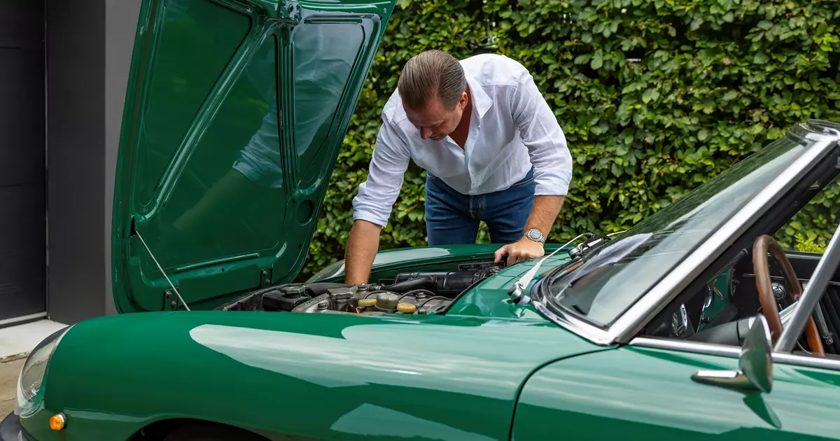 Billedet viser en mand i en hvid skjorte og jeans, som arbejder under motorhjelmen på en grøn klassisk bil. Bilen er parkeret foran en grøn hæk, og manden er fokuseret på at undersøge motoren. Interiøret i bilen afslører et træbeklædt rat, der tilføjer et retropræg.