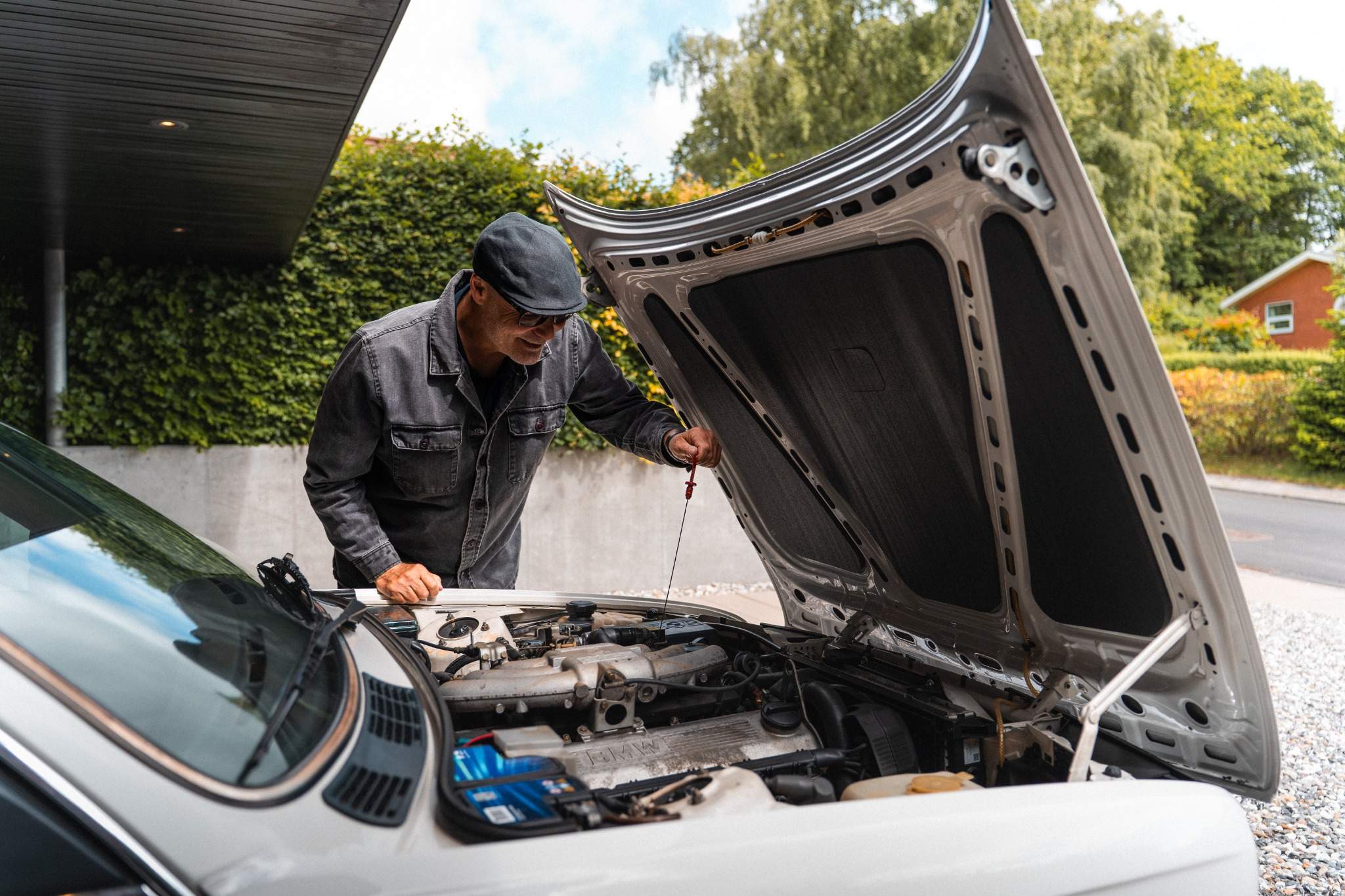 Billedet viser en mand i en hvid skjorte og jeans, som arbejder under motorhjelmen på en grøn klassisk bil. Bilen er parkeret foran en grøn hæk, og manden er fokuseret på at undersøge motoren. Interiøret i bilen afslører et træbeklædt rat, der tilføjer et retropræg.