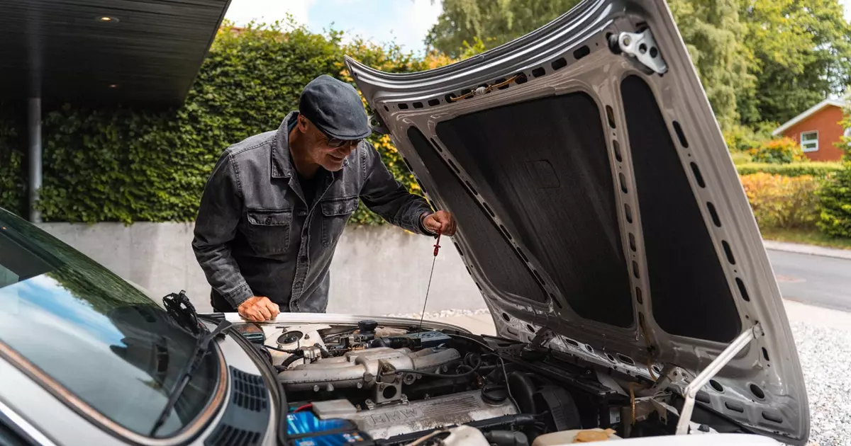 Billedet viser en mand i en hvid skjorte og jeans, som arbejder under motorhjelmen på en grøn klassisk bil. Bilen er parkeret foran en grøn hæk, og manden er fokuseret på at undersøge motoren. Interiøret i bilen afslører et træbeklædt rat, der tilføjer et retropræg.