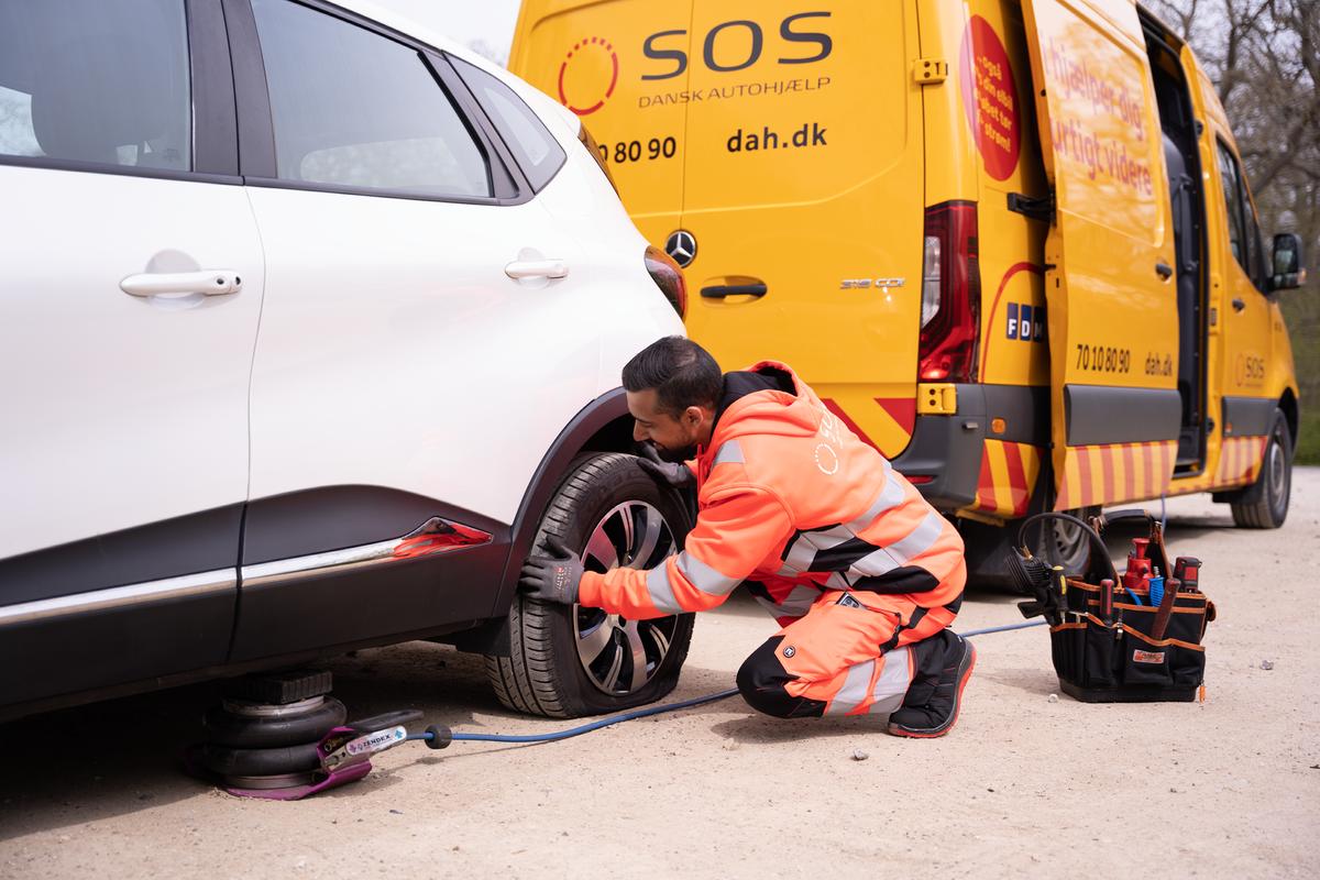 Billedet viser en mand i en hvid skjorte og jeans, som arbejder under motorhjelmen på en grøn klassisk bil. Bilen er parkeret foran en grøn hæk, og manden er fokuseret på at undersøge motoren. Interiøret i bilen afslører et træbeklædt rat, der tilføjer et retropræg.
