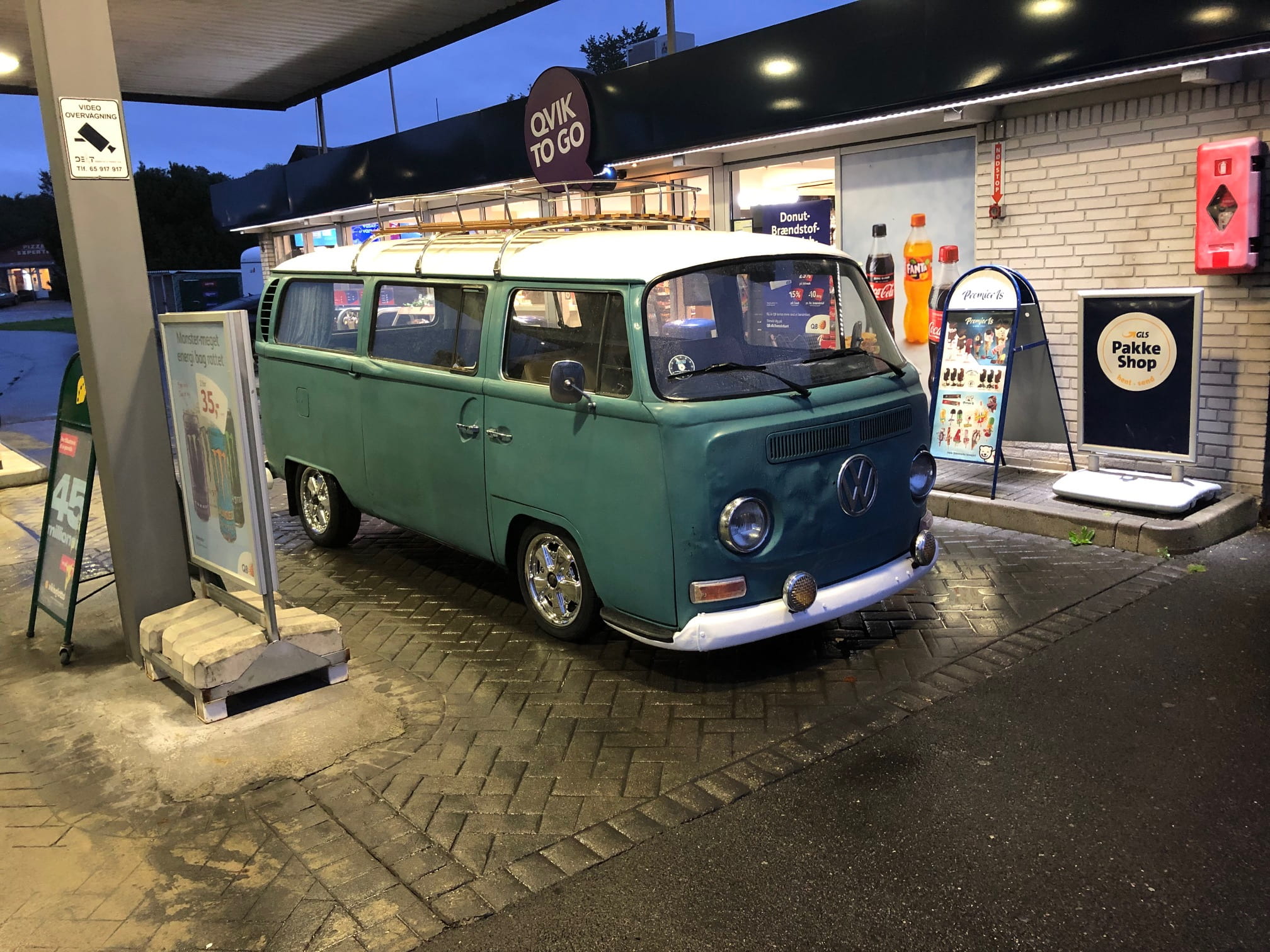 En klassisk grøn og hvid Volkswagen Type 2 (VW Bus) holder parkeret ved en Qvik To Go tankstation om aftenen. Bussen står under en overdækning med skiltning og reklamer for mad og drikke i baggrunden, mens området er vådt fra regn.