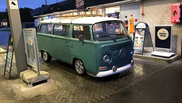En klassisk grøn og hvid Volkswagen Type 2 (VW Bus) holder parkeret ved en Qvik To Go tankstation om aftenen. Bussen står under en overdækning med skiltning og reklamer for mad og drikke i baggrunden, mens området er vådt fra regn.
