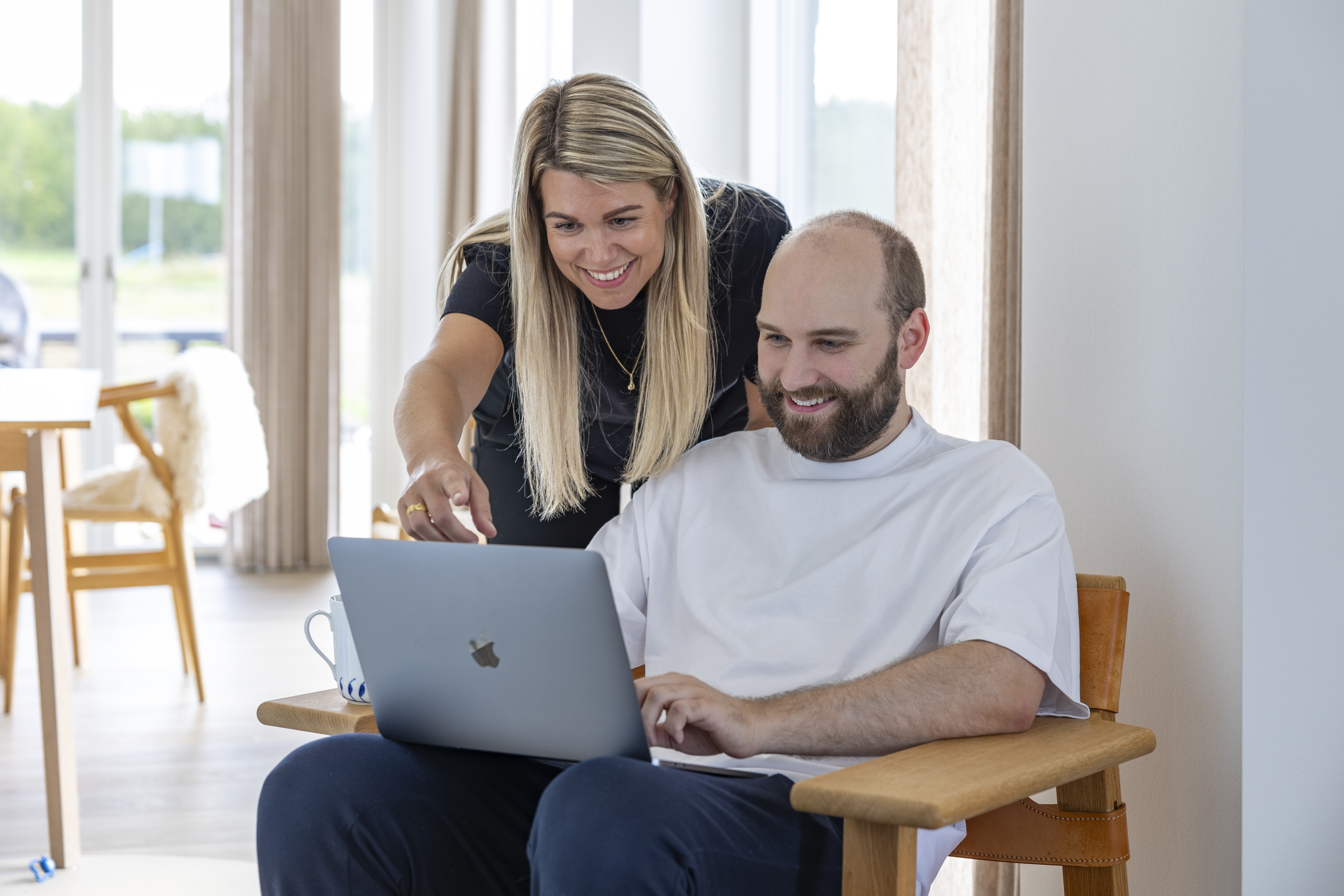 En mand sidder i en træstol med en bærbar computer, mens en kvinde står bagved og peger smilende på skærmen. Begge ser ud til at være engagerede i arbejdet. Rummet er lyst og indrettet med minimalistiske møbler.