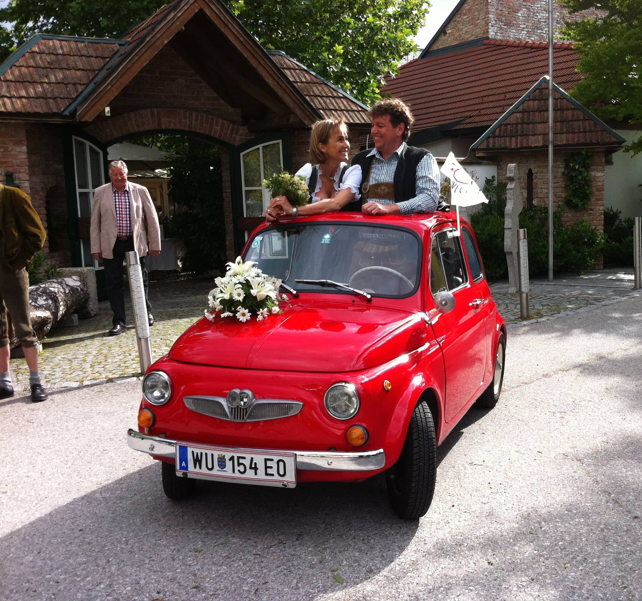 Et bryllupspar står i en lille, rød klassisk Fiat 500 dekoreret med hvide blomster på kølerhjelmen. Bruden holder en blomsterbuket og smiler mod gommen, der bærer traditionelt tøj. Nummerpladen viser "WU 154 EO". Baggrunden har et idyllisk hus med trædetaljer og en gårdsplads med brosten. En ældre mand står i baggrunden og ser på parret. Solen skinner, og stemningen er festlig.