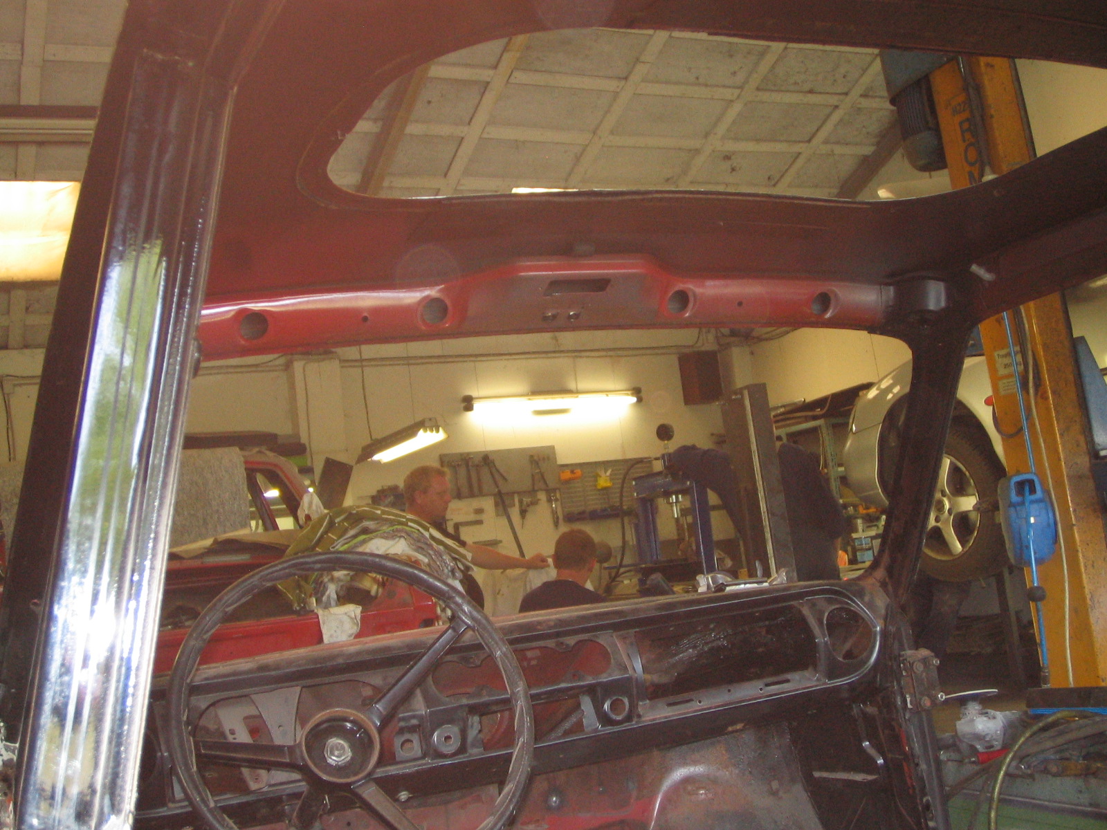 Udsigt gennem frontruden af en klassisk bil under restaurering. Rattet og instrumentpanelet er synligt, men delvist adskilt. I baggrunden er værkstedsområdet med flere personer i arbejde, værktøj og en hævet bil synlig. Loftet og belysningen skaber en industriel værkstedsstemning.