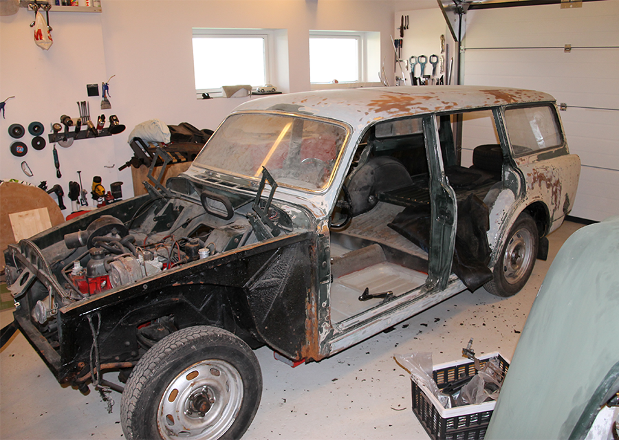 En gammel stationcar under restaurering i et værksted. Bilen mangler frontpaneler og døre, og karosseriet er rustent med afskallet maling. Inden i værkstedet er der værktøj, reservedele og en kurv med bildele på gulvet. Hylder langs væggen holder forskellige værktøjer og udstyr, mens to vinduer giver naturligt lys i rummet.