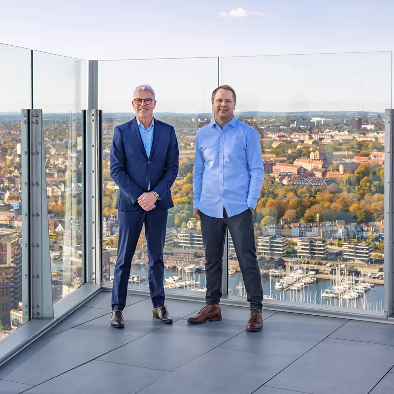 Billedet viser to mænd stående på en tagterrasse med glasafskærmning og en imponerende udsigt over en by. Den ene mand er iført en mørkeblå jakkesæt og sorte sko, mens den anden er mere afslappet klædt i en lys blå skjorte og mørke bukser. Bag dem ses en storby med boligblokke, marinaer og efterårsfarvede træer, hvilket indikerer en bynær placering med en blanding af natur og urban udvikling. Stemningen er professionel, og omgivelserne understreger højden og det spektakulære vue.