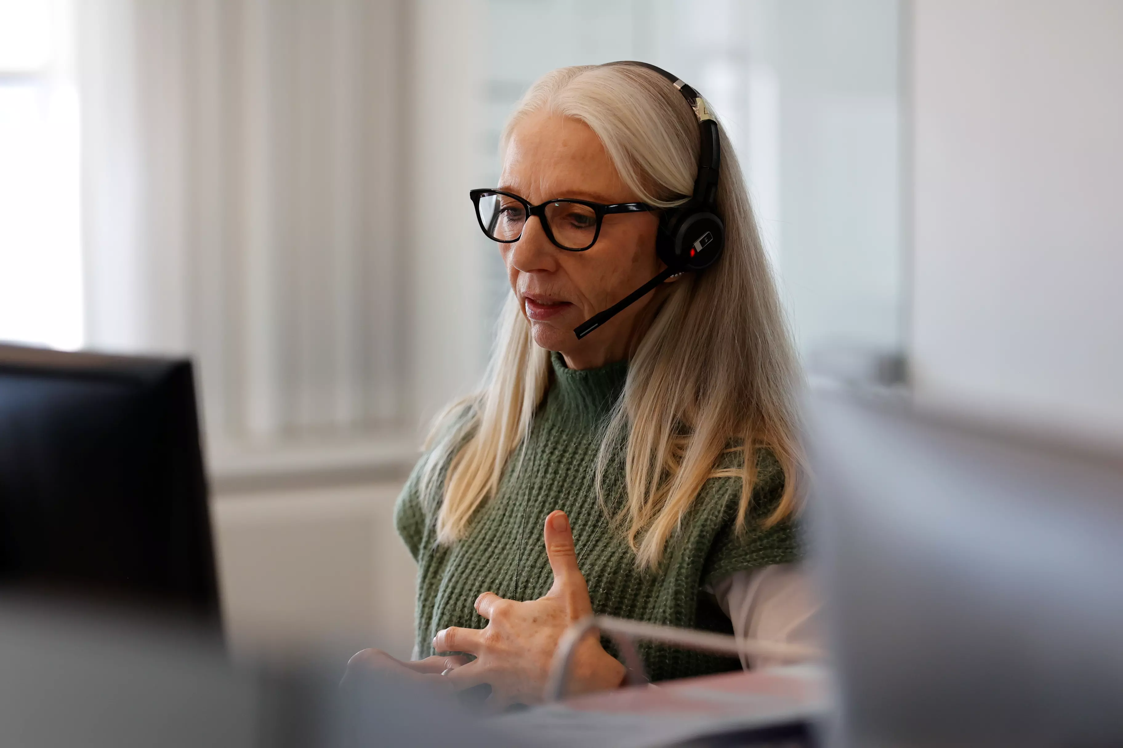 En kvinde med lyst hår og briller sidder foran en computer og taler i telefon. Hun har et venligt udtryk.