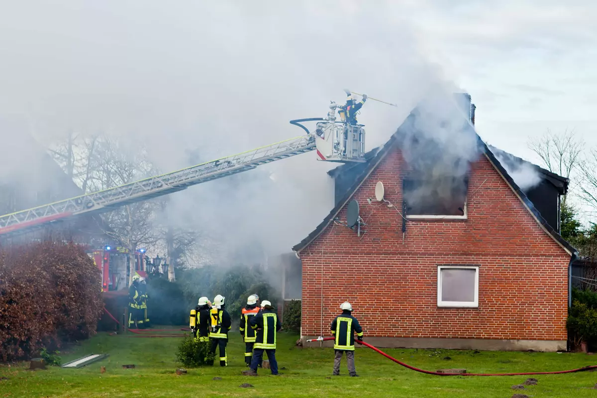 Billedet viser en alvorlig husbrand i en rød murstensbygning, hvor flammer og røg vælter ud fra taget og vinduerne. Brandfolk er til stede i fuld uniform og arbejder på at slukke branden. En stigevogn bruges til at nå de øverste dele af huset, mens andre brandfolk betjener brandslanger på jorden. Røgen fylder luften og skaber en dramatisk scene, der illustrerer indsatsen for at bekæmpe flammerne og redde ejendommen.