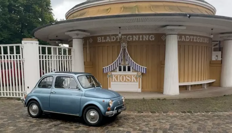 På billedet ses en blå veteranbil der holder parkeret på en brostensgade foran en bygning.