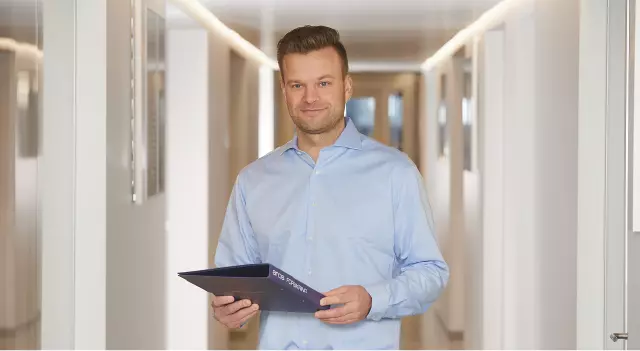 En mand iført en lyseblå skjorte står i en lys og moderne kontorgang. Han holder en mørk mappe med teksten 'Aros Forsikring' og smiler venligt til kameraet. Baggrunden viser en gang med døre og glaspartier, der skaber en professionel atmosfære.