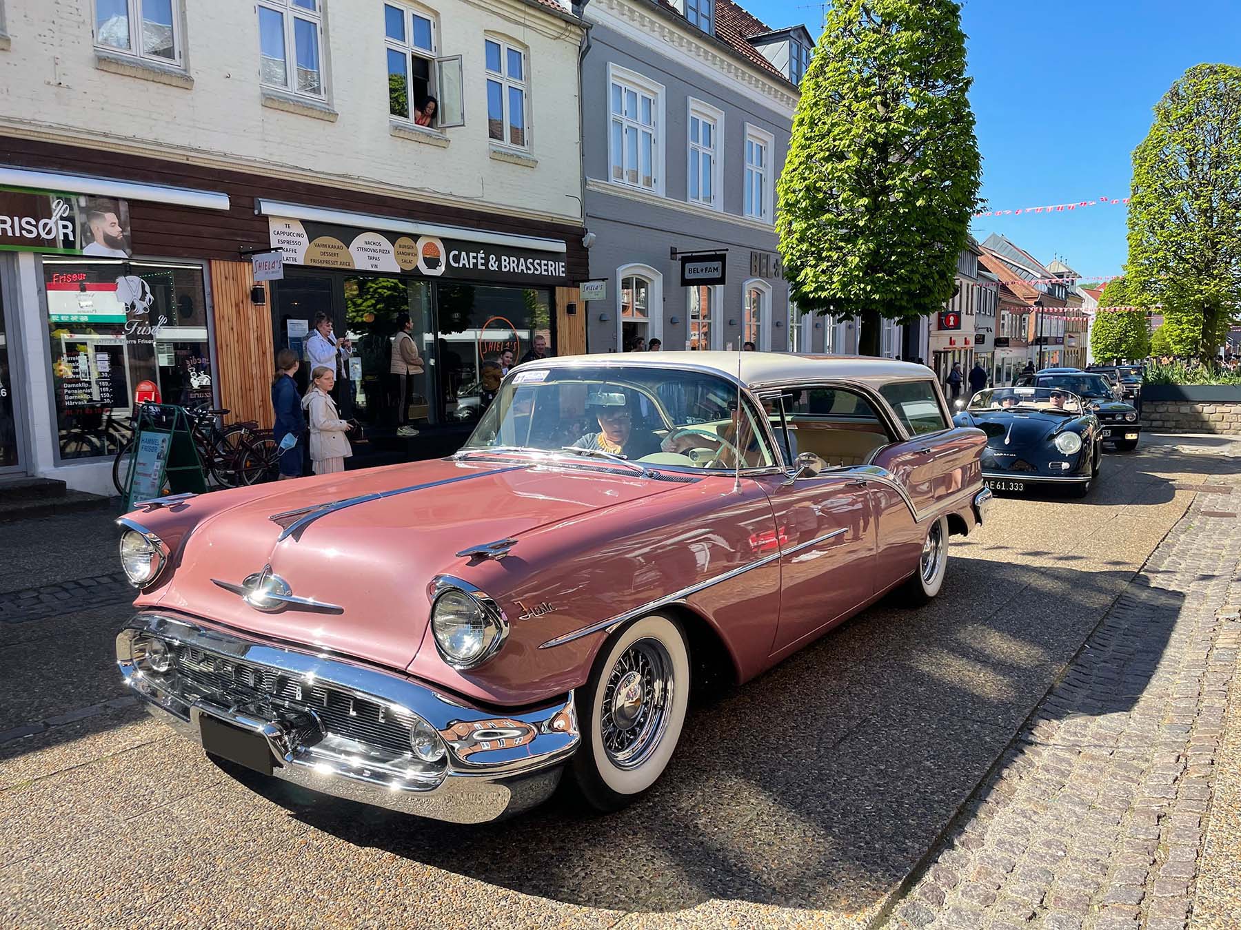 En smukt restaureret, gammel rosa klassisk bil med kromdetaljer holder parkeret på en brostensbelagt gade i solskin. Bilen har hvide væg-dæk og et skinnende udseende. I baggrunden er der flere klassiske biler på række og charmerende bygninger med cafeer og butikker, hvor mennesker står og nyder synet af bilerne.