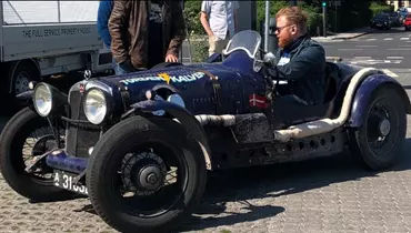 En mand med skæg og solbriller sidder i en mørkeblå, gammel racerbil med åbent cockpit og danske flag på siden. Bilen har et råt, vintage look med hvide udstødningsrør langs siden og påskriften "Torben Kalvø" i lyse farver. Et par personer står i baggrunden ved siden af en hvid varevogn, og solen skinner på en brolagt plads med skygger fra bilen og menneskerne.
