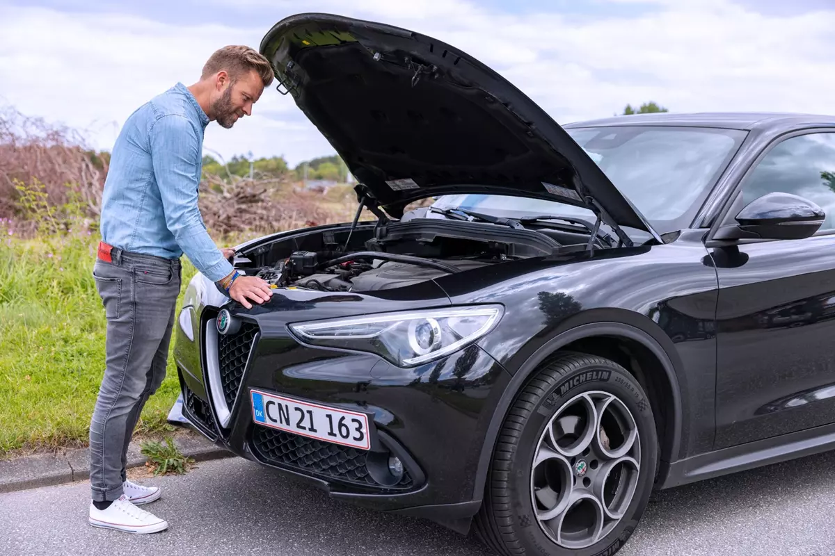 En mand i lysegrå jeans og en lyseblå denimskjorte står foran en sort Alfa Romeo med åben motorhjelm. Han undersøger motoren, mens bilen holder parkeret ved vejkanten.