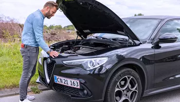 En mand i lysegrå jeans og en lyseblå denimskjorte står foran en sort Alfa Romeo med åben motorhjelm. Han undersøger motoren, mens bilen holder parkeret ved vejkanten.