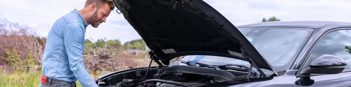 En mand i lysegrå jeans og en lyseblå denimskjorte står foran en sort Alfa Romeo med åben motorhjelm. Han undersøger motoren, mens bilen holder parkeret ved vejkanten.