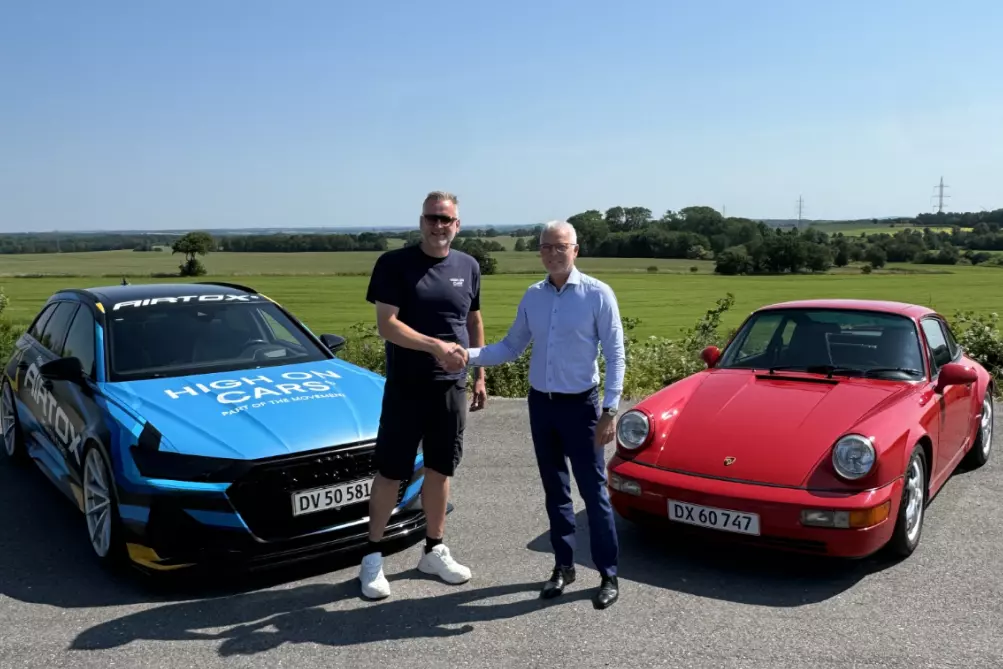 To mænd står på en solrig dag på en asfalteret vej mellem to biler – en blå bil med skriften "HIGH ON CARS" og en rød klassisk Porsche. De giver hinanden et håndtryk og smiler til kameraet. Bag dem ses åbne grønne marker og træer under en klar blå himmel. Den ene mand er iført en sort t-shirt og shorts, mens den anden har en lys skjorte og mørke bukser på.