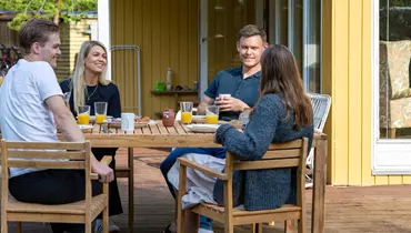 Fire personer sidder rundt om et træbord på en terrasse foran en gul træbygning. Bordet er dækket med glas appelsinjuice, kaffekopper og morgenmad. De smiler og ser ud til at nyde en afslappet samtale i solskinsvejr.