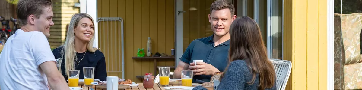 Fire personer sidder rundt om et træbord på en terrasse foran en gul træbygning. Bordet er dækket med glas appelsinjuice, kaffekopper og morgenmad. De smiler og ser ud til at nyde en afslappet samtale i solskinsvejr.